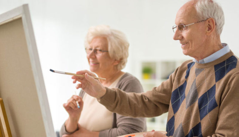 Older man and woman painting something on a canvas with paintbrushes at memory care facility