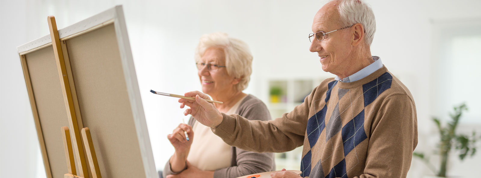 Older man and woman painting something on a canvas with paintbrushes at memory care facility